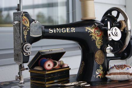 En äldre svart symaskin av märket Singer. En trälåda med sytråd och en nåldyna med nålar syns också.